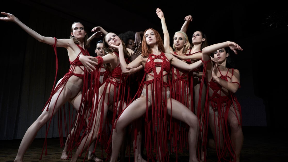 A group of dancers in red costumes striking dramatic poses with expressive arm movements on a dimly lit stage.