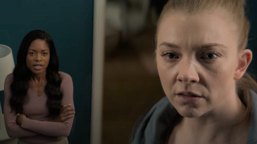 Two women are in a room; one stands in the background with arms crossed, and the other is in the foreground with a serious expression. The background woman wears a long-sleeved shirt; the foreground woman wears a hoodie.