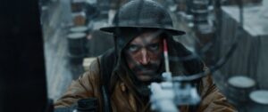 A person in muddy, rain-soaked attire and a helmet looks intently through a rain-splashed round frame in a dim, rustic setting with barrels in the background.