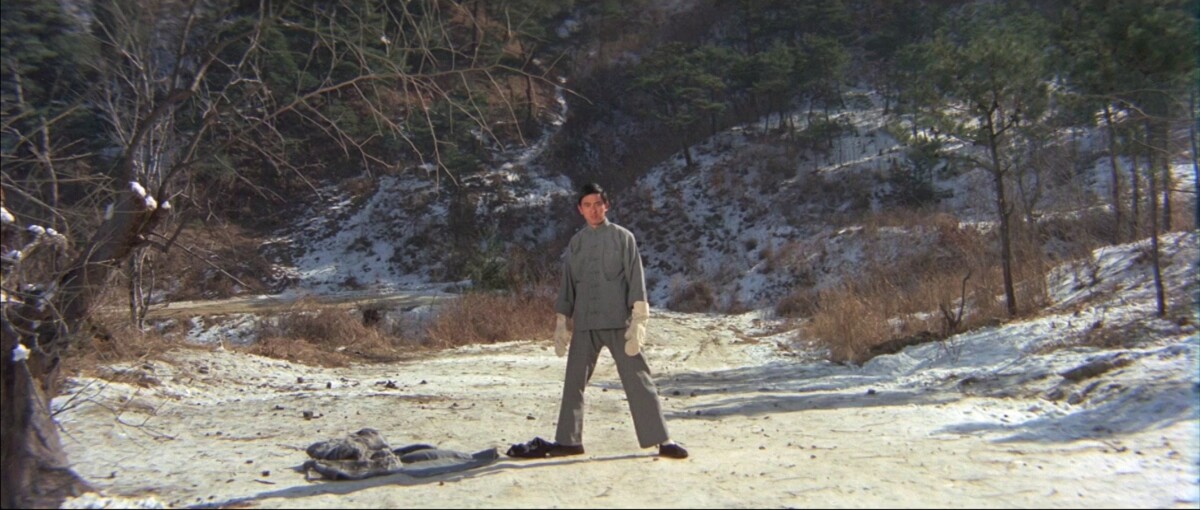 A person stands alone on a snowy path surrounded by trees and hills in winter, dressed in a gray outfit with a pair of black shoes and a scarf on the ground nearby.