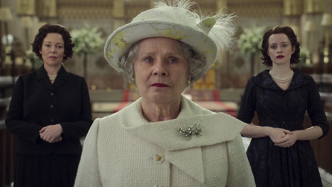Three women are dressed in period attire. The woman in the center wears a white hat and coat with a brooch, standing in a regal setting, while the other two women, in darker outfits, stand behind her.