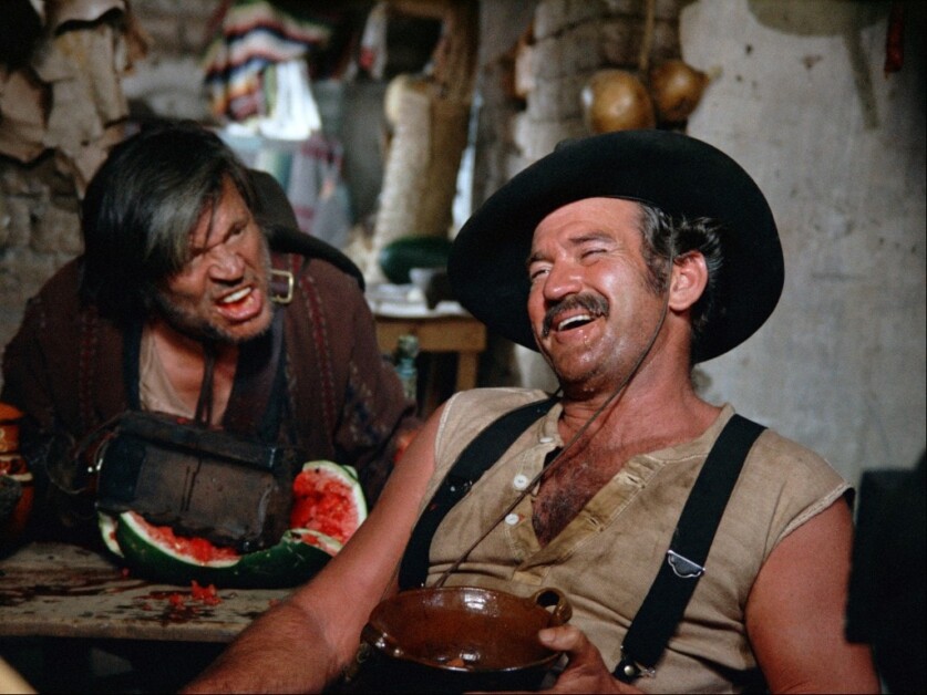 Two men are in a rustic interior: one man is leaning over with a watermelon, appearing angry, while the other, in a hat and suspenders, is laughing and holding a cup.