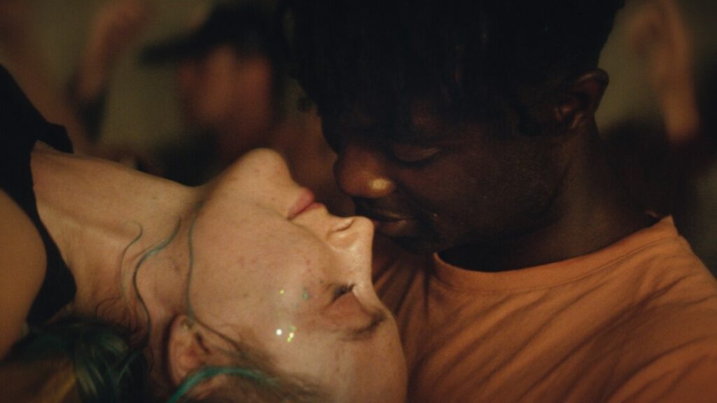 A person with glitter on their face is leaning back while another person brings their face close, both appearing to be at a social gathering.