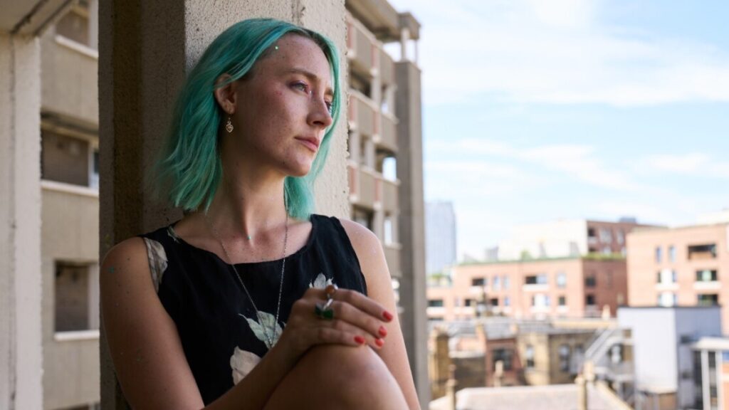 A person with blue hair and wearing a black sleeveless top sits outside on a balcony, gazing into the distance with a thoughtful expression. Urban buildings are visible in the background.