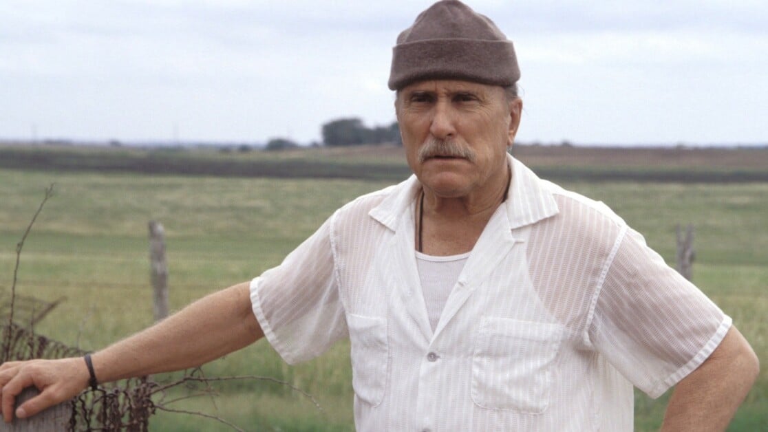 An older man with a mustache wearing a brown beanie, white shirt, and white undershirt, stands in a rural field with a fence and expansive landscape in the background.