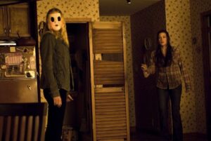 Two women are in a dimly lit room with floral wallpaper. One wears a mask and holds a weapon, while the other appears startled and is reaching out. A wooden door is partially open behind them.