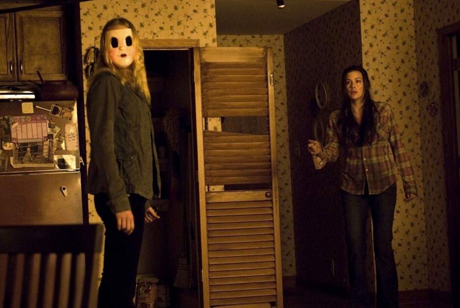 Two women are in a dimly lit room with floral wallpaper. One wears a mask and holds a weapon, while the other appears startled and is reaching out. A wooden door is partially open behind them.