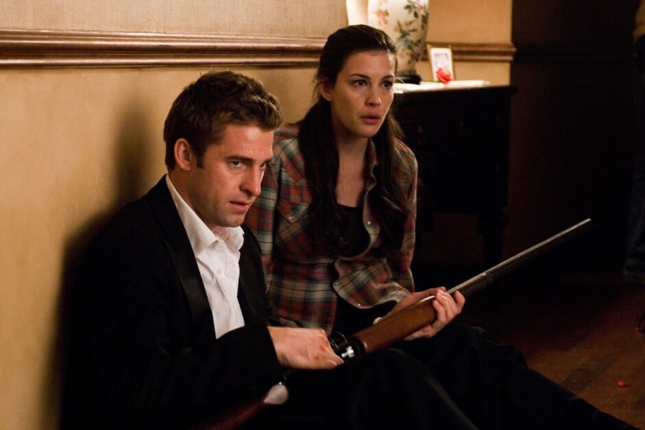 Two people sitting on the floor against a wall in a dimly lit room, with the man holding a rifle and the woman looking concerned.