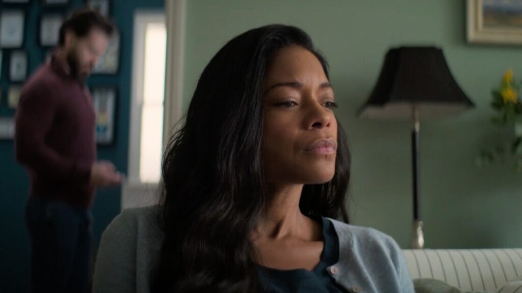 A woman with long dark hair sits indoors with a neutral expression, while a man in the background looks at his phone.