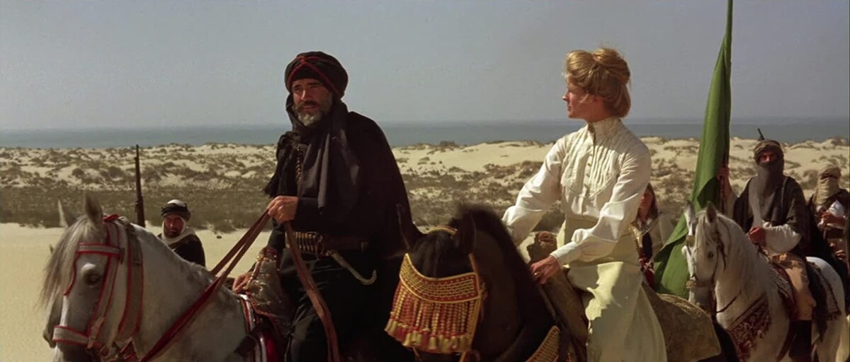 A man and a woman ride horses through a desert, accompanied by several others. The man wears a dark turban and cloak, while the woman is in a high-collared white dress.