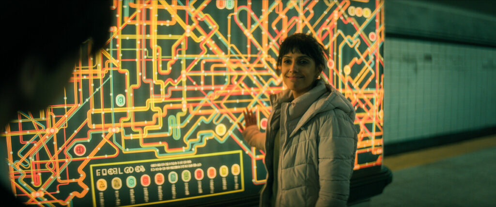 A woman in a white jacket stands beside an illuminated, intricate map in a possibly futuristic setting, smiling and pointing at the map. A blurred figure is visible in the foreground.