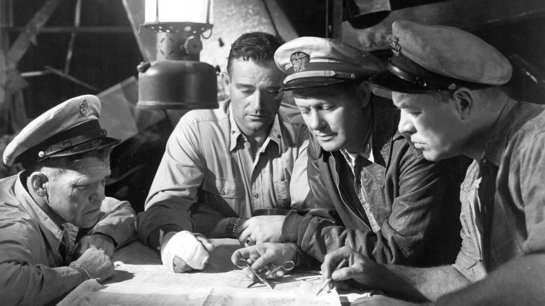 Four men in naval uniforms and another man are examining a map under a lantern light in a dimly lit room.