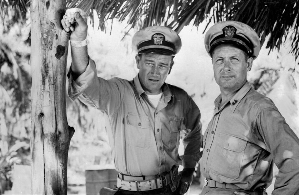 Two men in naval uniforms stand under a thatched roof. The man on the left leans with his arm on a post, while the man on the right stands with his hands on his hips.