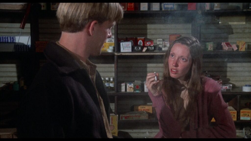 A man in a dark jacket and a woman in a pink coat holding a small item are talking in a store with shelves stocked with various products in the background.