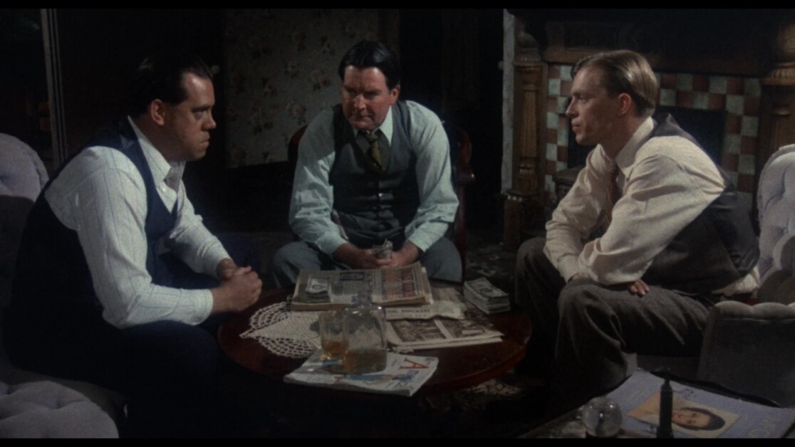 Three men in suits are seated around a table covered in papers, magazines, and a drink, engaged in a serious conversation in a dimly lit room.