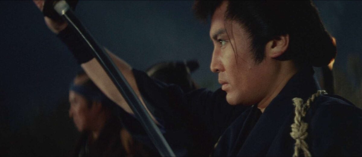 A man with a serious expression holds a raised sword in front of him, with another person in the background also raising a sword. The scene is set at night.