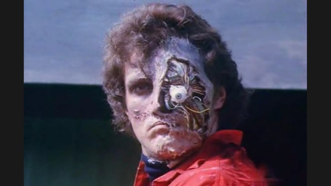 A person with visible cybernetic components on their partially damaged face wearing a red jacket.