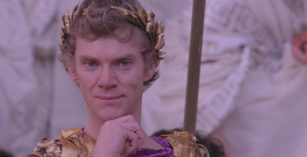 A person with blonde hair wearing a golden wreath and purple garment looks directly at the camera while resting their chin on their hand.