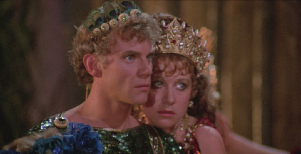 Two people in ornate costumes and crowns stand closely together, appearing serious with a dramatic background.