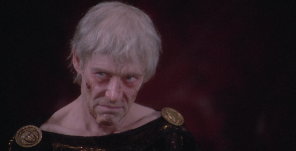 An older man with disheveled white hair and facial scars wears a dark garment with gold accents, looking sternly ahead.