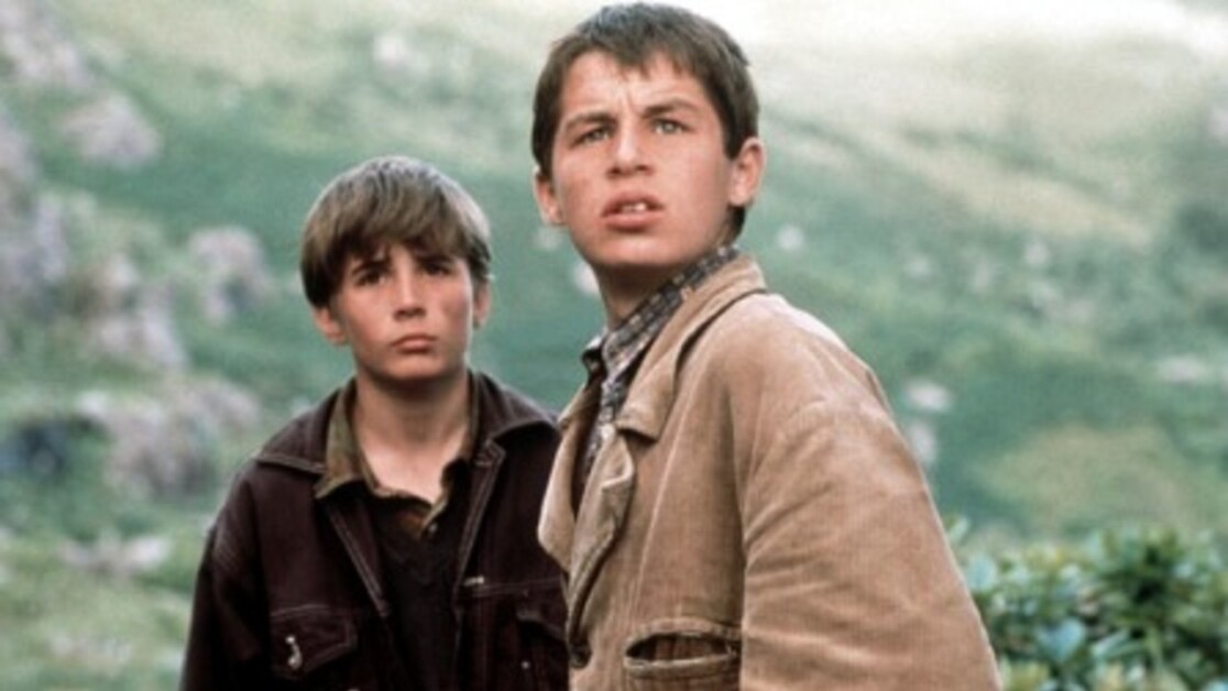 Two young boys are standing outdoors on a grassy hillside, both looking towards the distance. One boy is in a light brown jacket, and the other is in a dark jacket. The background is a hilly landscape.