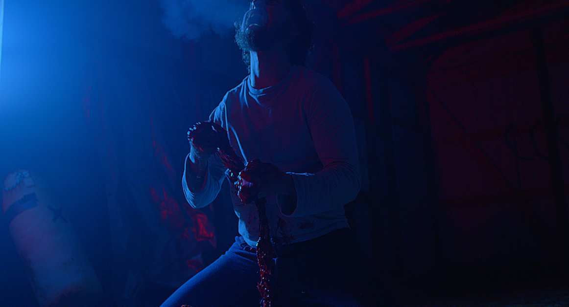 A person in a dimly lit room kneels and holds a bloodied object, with smoke visible near their head. The scene is illuminated by blue and red lighting.