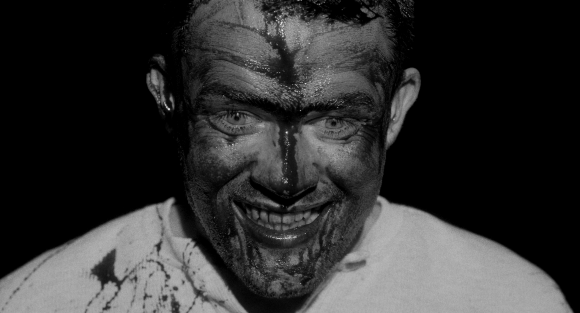 A man with a broad smile has his face and shirt splattered with a dark, liquid substance, standing against a dark background.