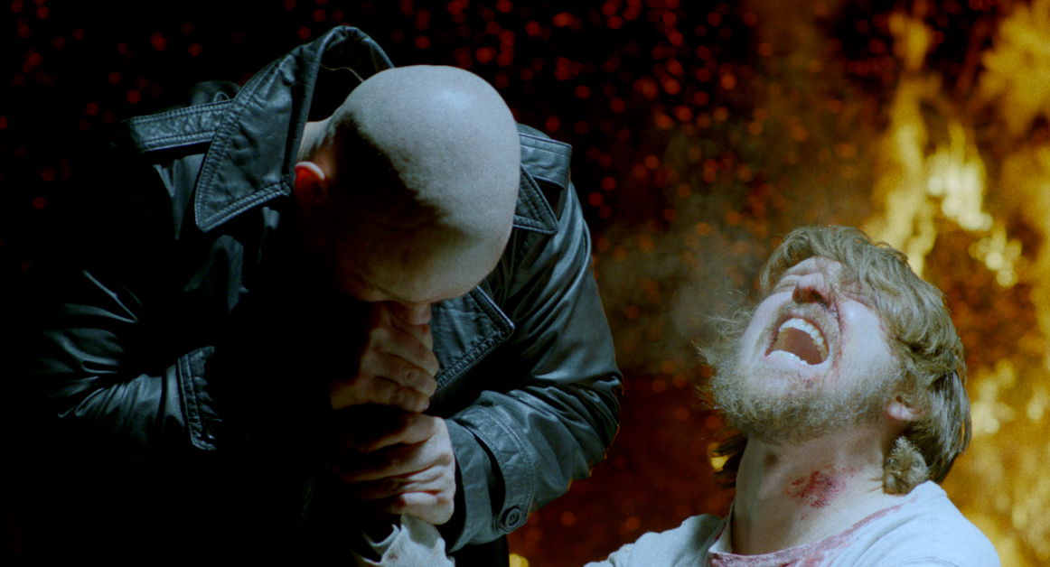 A bald man in a leather jacket bites his fist while kneeling next to a man with blood on his neck who is screaming in front of a background of flames.