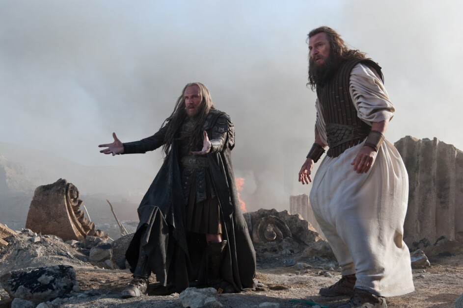 Two men in medieval-style clothing stand in a desolate, smoky landscape. One man gestures with open hands while the other looks forward with a serious expression. Ruins and fire can be seen behind them.
