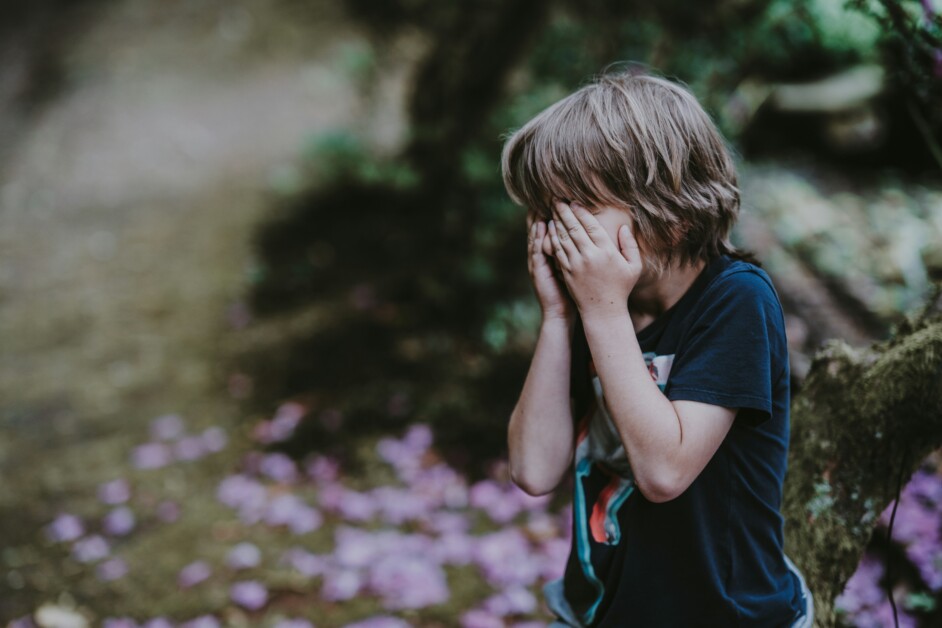 A child with blond hair, wearing a dark blue T-shirt, covers their face with both hands while standing outdoors near a tree and scattered petals.