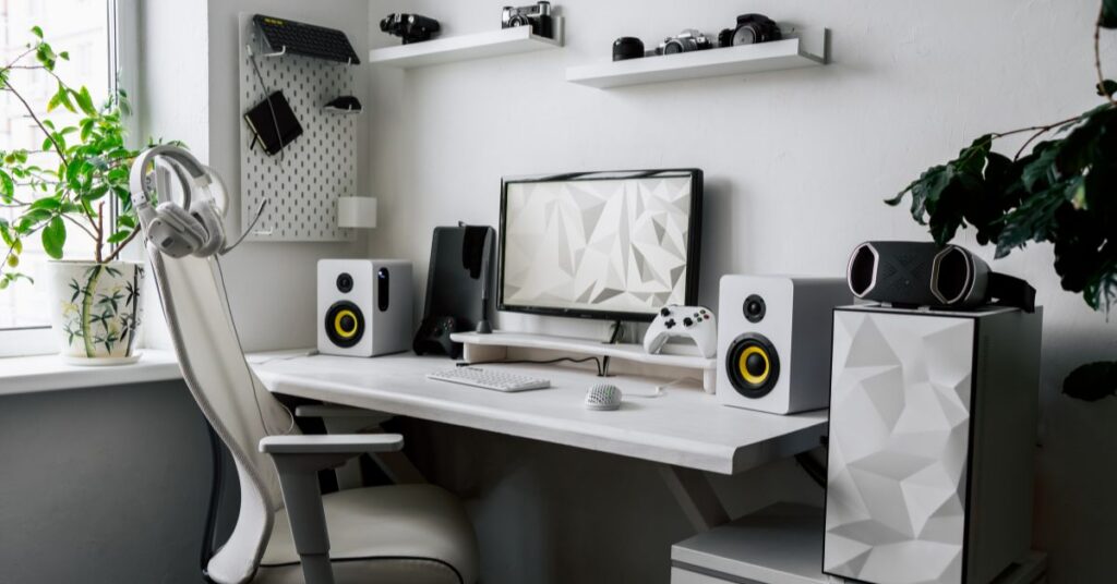 A modern home office setup with a white desk, computer monitor, keyboard, two speakers, a gaming controller, headphones, and decorative plants. Shelves with cameras are mounted on the wall above.