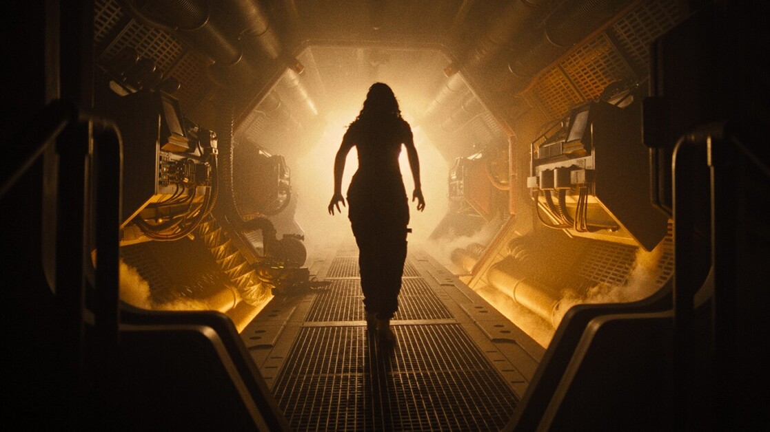 A person walks through a dimly lit, industrial corridor with machinery lining the walls and light shining from behind.