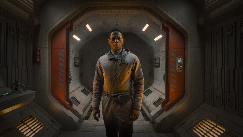 A person in a space suit stands in a futuristic corridor with red and white lighting. Control panels and a fire extinguisher are visible on the walls.