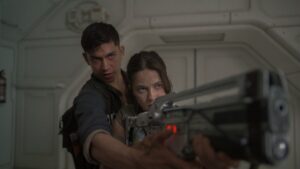 Two people in a futuristic setting hold and aim a large, high-tech weapon together, with a tense and focused expression.