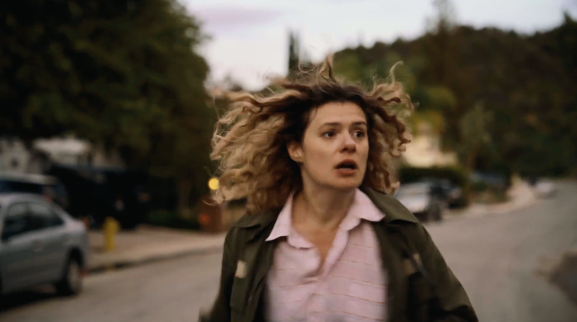 A woman with curly hair wearing a green jacket and a pink shirt is running down a street with a worried expression. Trees and cars line the street in the background.