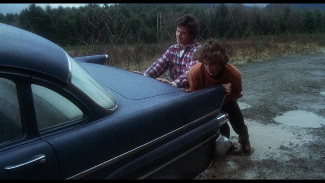 Two individuals are pushing a blue vintage car on a muddy, rural road surrounded by trees.