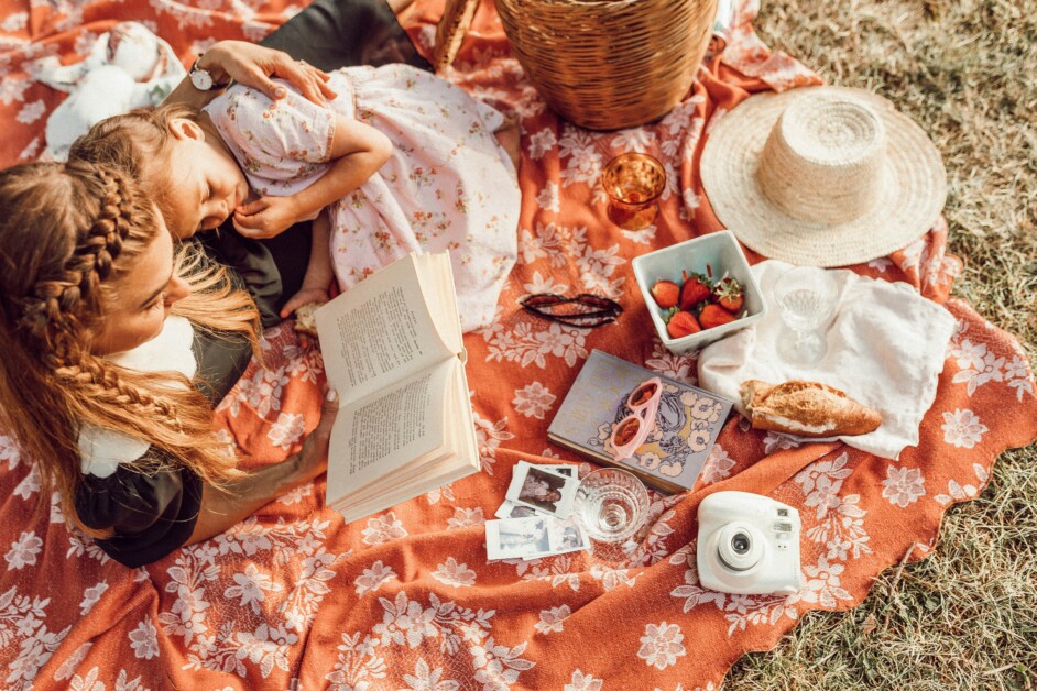 Two people, one reading a book and the other resting on a picnic blanket. The blanket has various items including strawberries, glasses, a hat, a camera, photos, and a croissant.