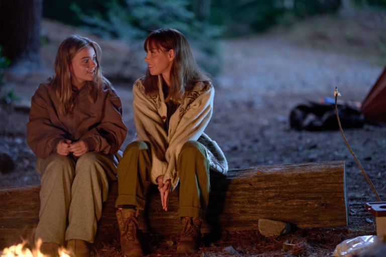 Two people sit on a log by a campfire in a forested area at dusk, engaged in conversation. One is in a brown sweater and sweatpants, the other in a beige sweater and green pants.