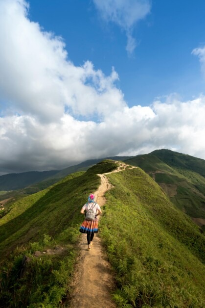 A person in traditional clothing walks along a narrow, winding path atop a lush green hill with a clear blue sky and scattered clouds in the background.