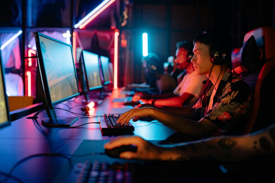 Several individuals wearing headsets are seated in a dimly lit room with neon lights, intensely focused on playing video games on desktop computers.