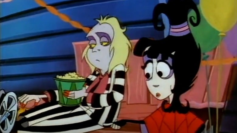 Two animated characters, one with messy blonde hair in a striped suit eating popcorn, and the other with black hair styled upwards, sitting together indoors with balloons in the background.
