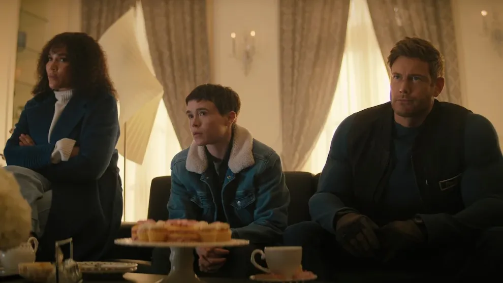 Three people sit on a couch in a well-lit room with white curtains, facing forward. In front of them is a table with teacups and a plate of pastries.