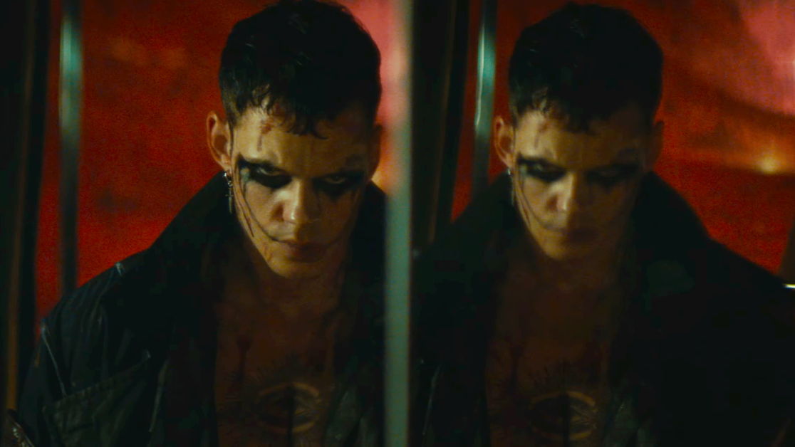 A person with dark makeup around their eyes is reflected in a mirror under dim, dramatic lighting.