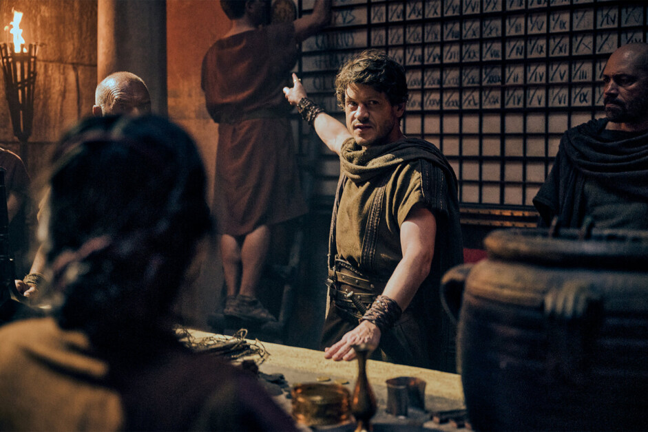 A person in period costume points to a grid on the wall while others in similar attire look on. The setting appears historical with dim lighting and props.