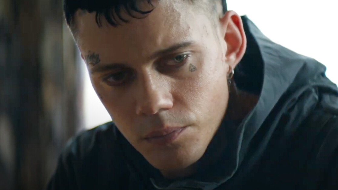 A person with a teardrop face tattoo and multiple piercings is shown in a close-up, looking downward with a serious expression. They are wearing a dark hoodie.