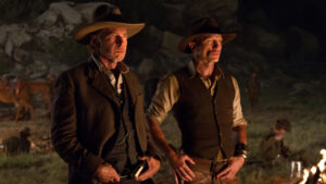 Two men in Western attire and hats stand outdoors at night, with other people and horses visible in the background.