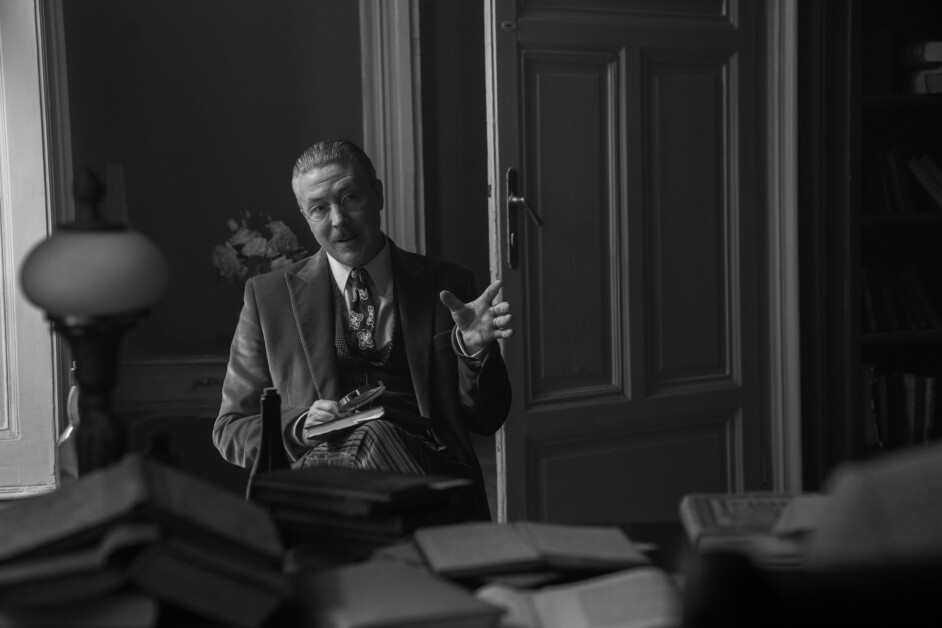 A man in a suit sits in a dimly lit room, holding a notebook and making a hand gesture. The room has a table with scattered books, a lamp, and a partially open door in the background.