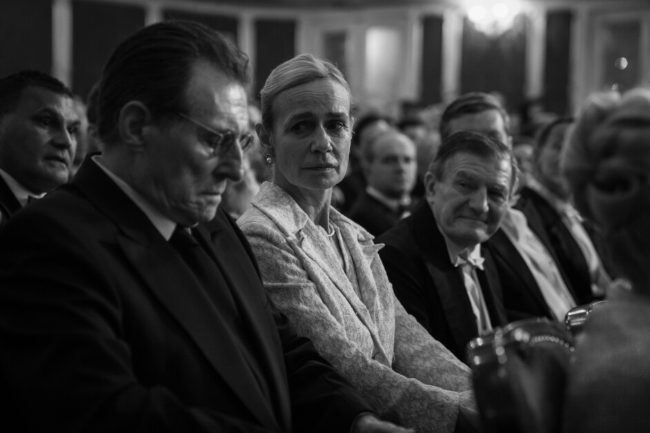 Black and white image of a formal audience. A woman in a light-colored jacket looks intently at a man wearing glasses who is looking down. Other people in formal attire are seated around them.