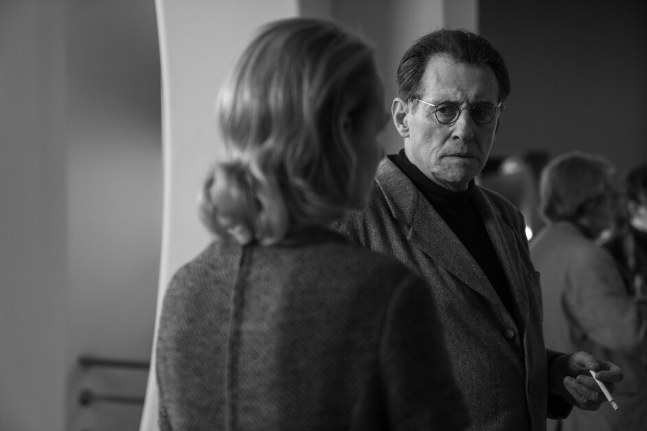 A black-and-white image of two people in conversation; one person, seen from behind, faces another who is wearing glasses and a suit, holding an object in hand.