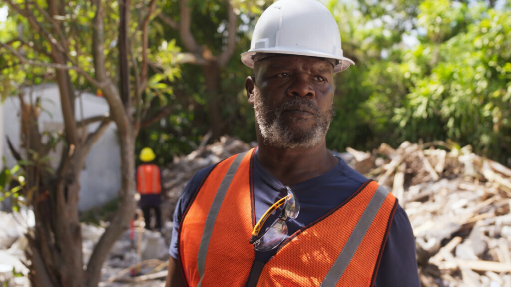 Man in an orange safety vest and white hard hat stands at a construction site, with another worker and debris in the background.